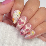 Close-up of a hand with pink glittery nails featuring floral and heart designs on a white background.