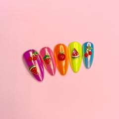 Five almond-shaped press-on nails with glossy metallic colors and fruit designs, arranged on a solid pink background. The nails feature pink, magenta, orange, lime green, and blue shades, decorated with red cherries, strawberries, and watermelon slices.